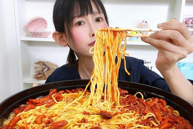 "大量の辛そうな麺料理を箸で持ち上げる女性。白い棚とインテリアが背景にある室内。"