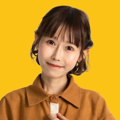 "おごせ綾さんの近影。黄色の背景に茶色の服を着て笑顔を見せる姿" 