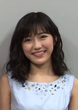 渡辺麻友さんが笑顔で正面を見ている上半身のポートレート写真です。黒髪のボブスタイルで、水色の襟元にビーズ飾りのついた衣装を着ています。背景は無地の壁です。