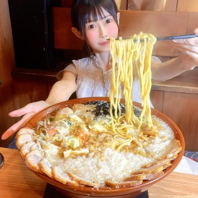 "大きなラーメンどんぶりを持ち上げた麺とともに見せる女性。店内の木製テーブルと背景が写っている。"