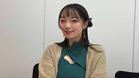 ベージュのカーディガンと緑のトップスを着た女性が椅子に座り、笑顔でカメラを見つめている室内の様子。