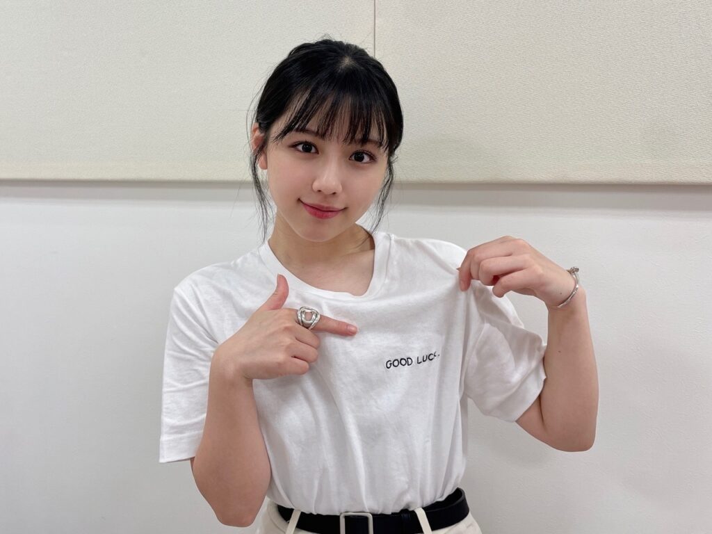"白いTシャツに『GOOD LUCK.』と書かれた文字を指さす若い女性。笑顔でカメラを見つめている。