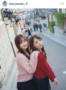 森香澄と女性が、石畳の道を背景に並んでポーズをとっている。森香澄は赤色の服を着て振り返り、隣の女性はピンク色の服でカメラを見ている。