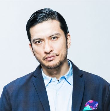 白い背景で撮影された、アジア人男性のポートレートです。男性は、濃い紺色のジャケットと水色のシャツを着用し、胸ポケットにはカラフルなポケットチーフが見えます。カメラをまっすぐ見つめており、真剣な表情をしています。