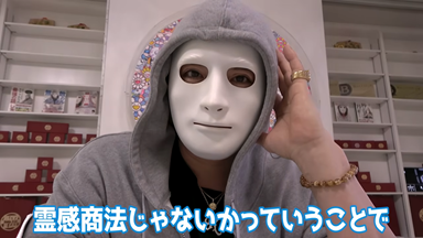  白い仮面をつけグレーのパーカーのフードをかぶったYouTuberラファエルが、商品を陳列した棚を背景に話しており、画面下部には「霊感商法じゃないかっていうことで」というテロップが表示されている。