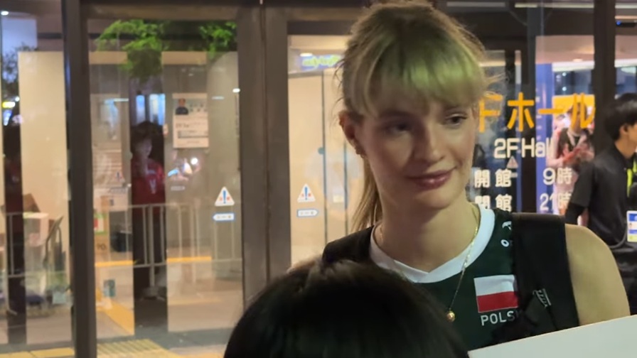 この写真は、バレーボールの試合後、会場の外でファンに対応している場面だと思われます。ピアセッカ選手はポーランド代表のユニフォームを着用しており、ファンと交流している様子がうかがえます。後ろには「2F Hall」といった案内表示が見え、夜間の建物入口付近で撮影されたことが分かります。