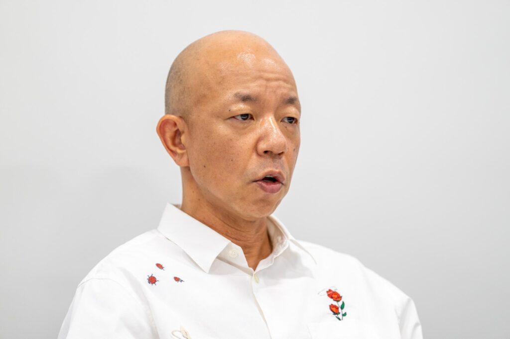 白いシャツを着た、スキンヘッドの男性が口を開けて話しています。