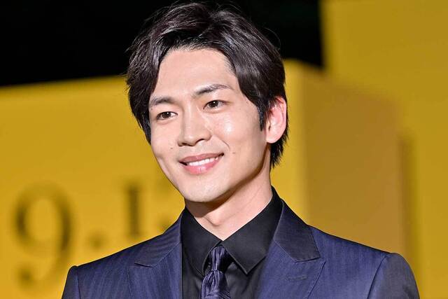 黒いスーツとネクタイを着用した、笑顔の男性のバストアップ写真です。メディアのイベントか何かの席で撮影されたポートレートのように見受けられます。