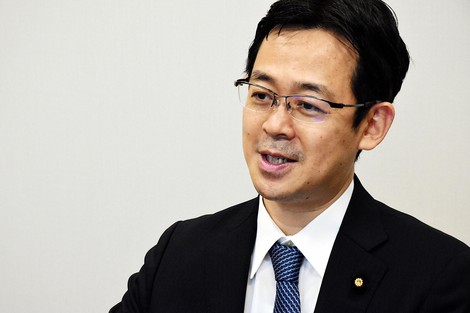 スーツとネクタイを着用し、メガネをかけた男性のバストアップ画像です。口元には微笑みを浮かべています。