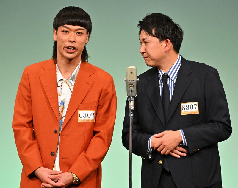 M-1グランプリの審査員席に座るお笑いコンビ「バッテリィズ」の二人が、真剣な表情で審査している様子です。
