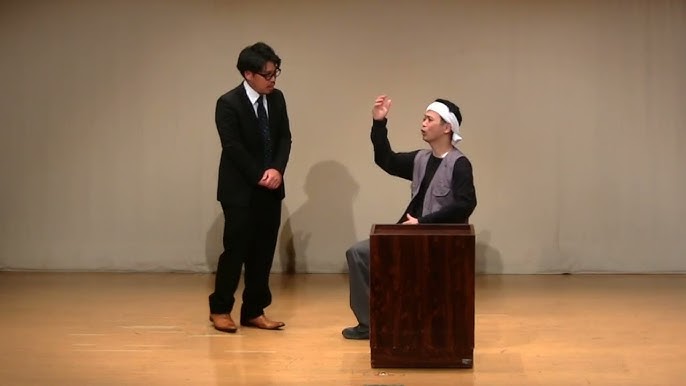 拍手をしているスーツ姿の男性と、隣に立つジャケット姿の男性のお笑いコンビの舞台の様子