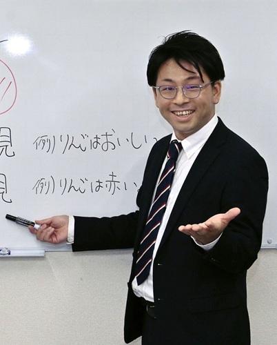 白いホワイトボードの前で、スーツを着た男性の塾講師が笑顔で話しています。ホワイトボードには手書きで文字が書かれています。