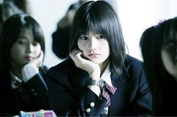 教室で制服を着た女子生徒が、少し憂鬱そうな表情で頬に手を当てて座っている様子。