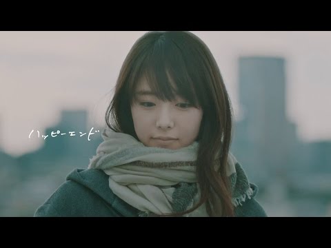 「厚手のマフラーを巻いたロングヘアの女性が、冬の都市の風景を背景に、うつむき加減で物憂げな表情を浮かべているミュージックビデオの一場面です。」