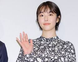 「白黒の柄の服を着たショートヘアの女性が、会見のような場所で穏やかに微笑みながら右手を振っている上半身の写真です。」