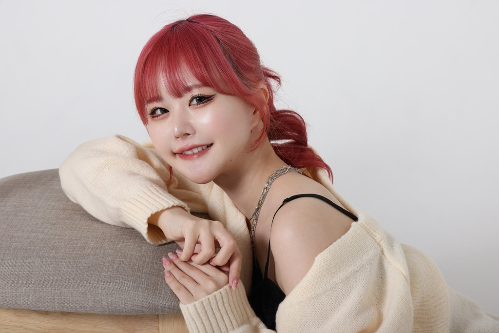 "鮮やかな赤い髪の整形アイドル轟ちゃんが、オフホワイトのカーディガンを羽織り、クッションに寄りかかりながらカメラに優しく微笑んでいます。"