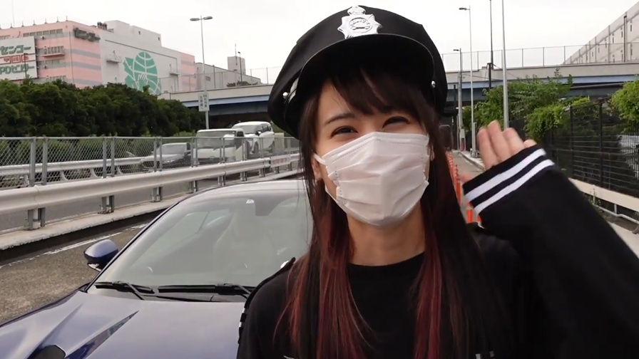 フェラーリの前に立ち、警察官の帽子を被り、白いマスクをつけ、片手を上げて挨拶している女性。
