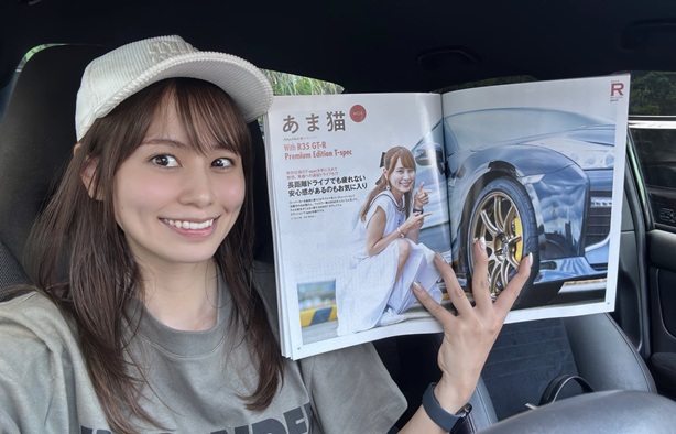 車の助手席に座ってカメラに笑顔を向けるあま猫さんが、自身の顔と高級車「NISSAN GT-R」が掲載された雑誌を広げて見せている。