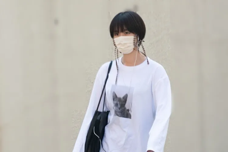 「猫がプリントされた白いTシャツを着たボブヘアの女性が、マスクをしてイヤホンを耳にかけ、街なかを歩いている様子の写真です。」