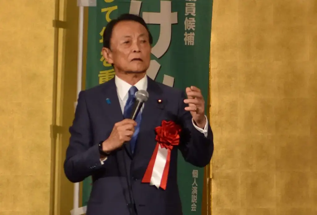 自由民主党副総裁の麻生太郎氏が、選挙イベントまたは個人演説会でマイクを持ってスピーチしている様子。胸には赤いリボンを着用している。