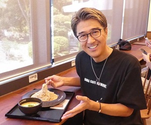 窓際のカウンター席で、眼鏡をかけた中年男性が笑顔で蕎麦のような料理を食べている。彼は黒いTシャツを着ており、リラックスした様子でカメラに視線を向けている。