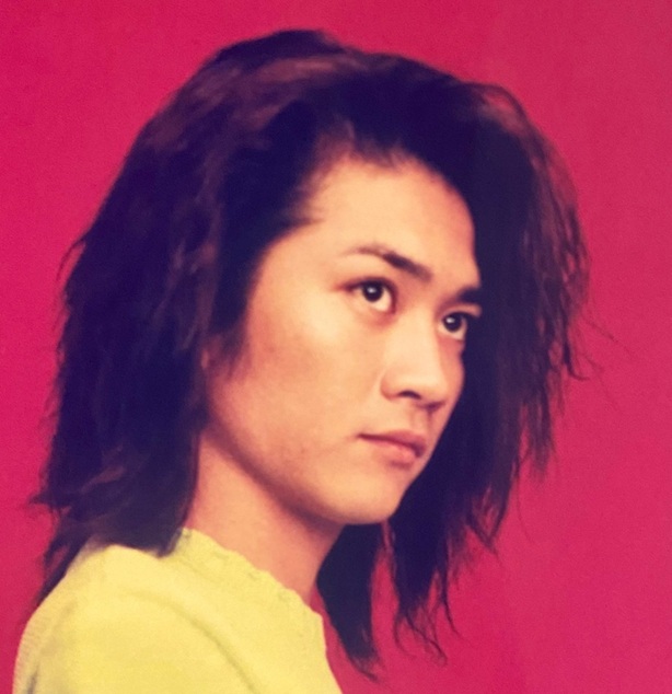 赤い背景の前で、横向きに鋭い眼差しを向ける長髪時代の若き国分太一さんの写真